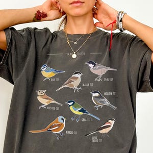 May include: Dark gray t-shirt featuring a detailed illustration of various bird species, including Blue Tit, Marsh Tit, and Coal Tit, with their names printed above. The shirt has a relaxed fit and the word "PEPPER" is printed on the bottom left.