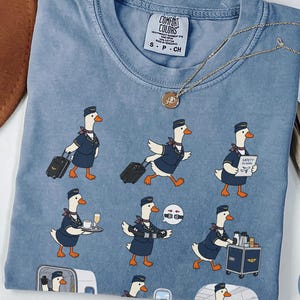 May include: A blue jean coloured t-shirt featuring a graphic print of cartoon geese dressed as flight attendants performing various tasks. The shirt has a crew neckline and a Comfort Colors brand label on the inside collar showing size S-CH.