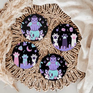 May include: Set of four round coasters with a spooky theme. Each coaster features a black background with a cat, skull, and paw prints in pastel colors. The coasters are displayed on a woven placemat.