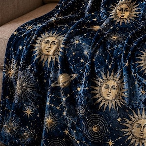 Peut inclure: Un plaid en peluche bleu foncé orné d'un motif céleste composé d'illustrations dorées de soleils avec des visages, de croissants de lune, de planètes et de diverses constellations d'étoiles réparties sur toute la surface du tissu.