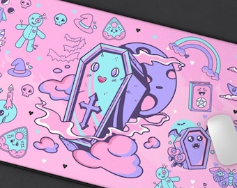 May include: A pink rectangular mousepad featuring a cartoon design with a coffin, moon, cat, and other spooky elements. The design includes a rainbow, clouds, and various whimsical illustrations in pastel colors.