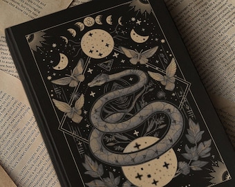 Può includere: Un diario con copertina rigida nera con un'illustrazione dettagliata di un serpente avvolto attorno a una luna crescente e stelle. Il diario è circondato da vecchi libri, una candela accesa e altri oggetti vintage.