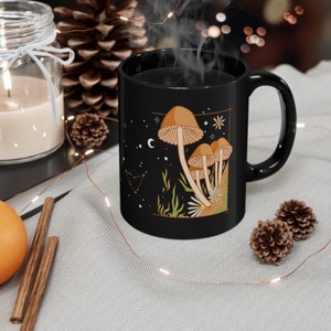 May include: Black ceramic mug featuring a whimsical mushroom design with a celestial theme. The illustration includes brown mushrooms, stars, a crescent moon, and a constellation. The mug has a black handle and is filled with a steaming dark liquid.