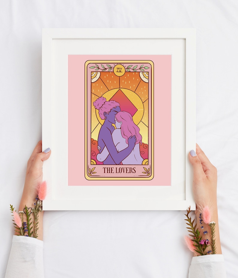Op de afbeelding: Een ingelijste print van een tarotkaart, "De Geliefden". Het kunstwerk toont twee figuren die elkaar omarmen, weergegeven in tinten paars, roze en goud. De kaart staat tegen een roze achtergrond, omlijst door een witte rand.