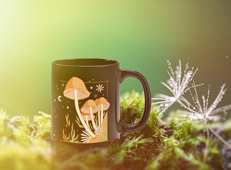 May include: Black ceramic mug featuring a whimsical illustration of mushrooms with a celestial design. The artwork includes tan and orange mushrooms, stars, and a crescent moon. The mug has a black handle and is set against a mossy background.