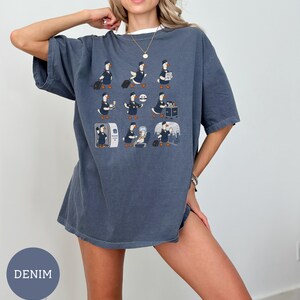 May include: A denim-coloured oversized t-shirt featuring a graphic print of nine cartoon ducks dressed as flight attendants performing various tasks like pulling luggage, serving drinks, and assisting passengers on an aeroplane in a grid layout.