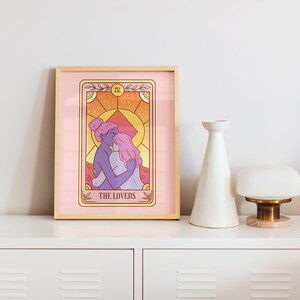 Op de afbeelding: Een ingelijste tarotkaartafdruk met een illustratie van twee figuren die elkaar omarmen, met de tekst "THE LOVERS". Het kunstwerk heeft een roze en oranje kleurenschema en wordt gepresenteerd op een witte ladekast.