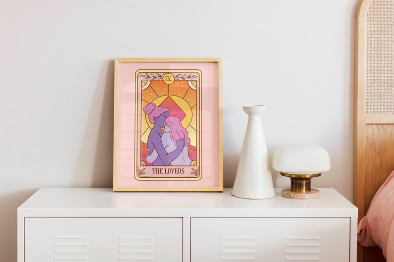 Op de afbeelding: Een ingelijste tarotkaartafdruk met een illustratie van twee figuren die elkaar omarmen, met de tekst "THE LOVERS". Het kunstwerk heeft een roze en oranje kleurenschema en wordt gepresenteerd op een witte ladekast.
