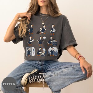 May include: A charcoal grey short-sleeve t-shirt featuring a graphic print of nine cartoon ducks dressed as flight attendants in navy blue uniforms. The ducks are depicted performing various tasks like pulling luggage, serving drinks, and greeting passengers.