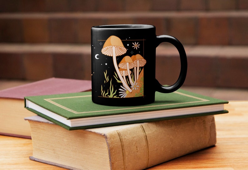 May include: Black ceramic mug featuring a whimsical illustration of orange and tan mushrooms against a starry night sky with a crescent moon and stars. The mug is placed on a stack of books with green and brown covers.