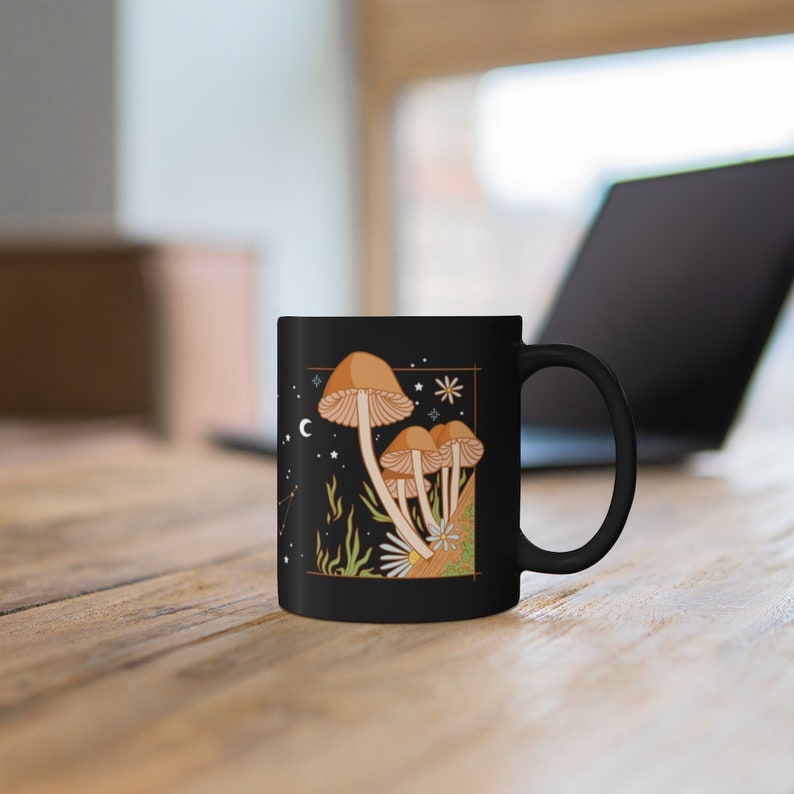 May include: Black ceramic mug featuring a whimsical illustration of mushrooms with a celestial design. The artwork includes tan mushrooms, stars, a crescent moon, and green foliage against a black background. The mug is on a wooden surface.