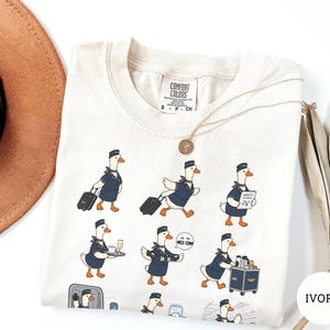 May include: An ivory t-shirt featuring a graphic print of multiple cartoon geese dressed as flight attendants in navy blue uniforms. The geese are depicted performing various tasks like pulling luggage, serving drinks, and pushing a service cart.