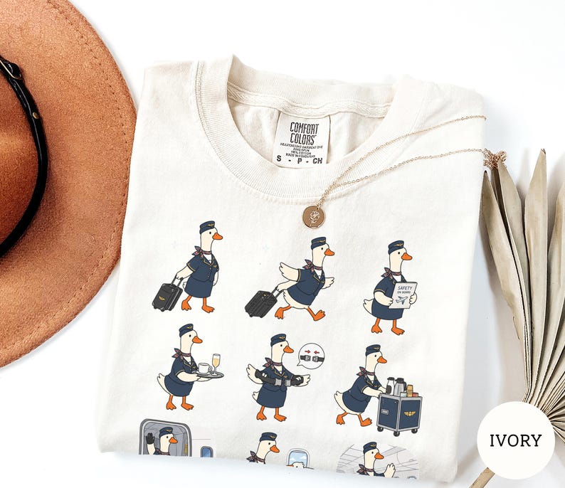 May include: An ivory t-shirt featuring a graphic print of multiple cartoon geese dressed as flight attendants in navy blue uniforms. The geese are depicted performing various tasks like pulling luggage, serving drinks, and pushing a service cart.