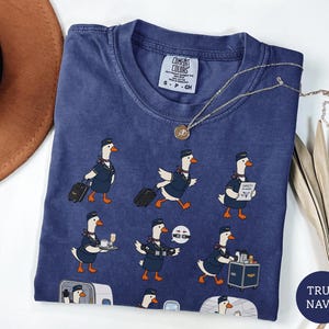 May include: A navy blue short-sleeve t-shirt featuring a repeating graphic print of white geese dressed as flight attendants. The geese are depicted performing various tasks like pulling luggage, serving drinks, and demonstrating safety procedures.