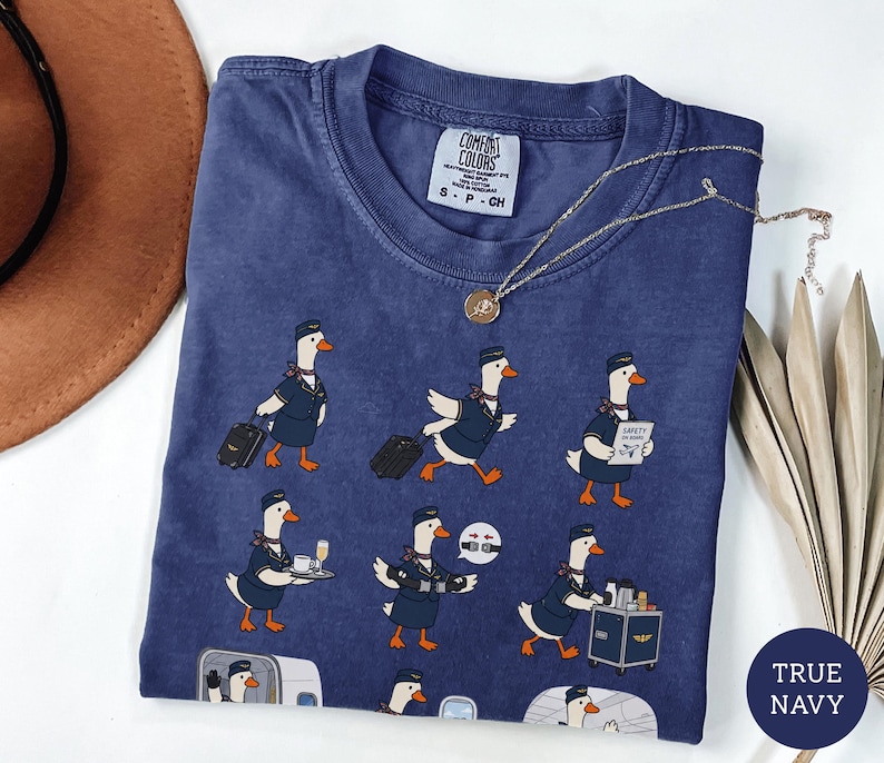 May include: A navy blue short-sleeve t-shirt featuring a repeating graphic print of white geese dressed as flight attendants. The geese are depicted performing various tasks like pulling luggage, serving drinks, and demonstrating safety procedures.