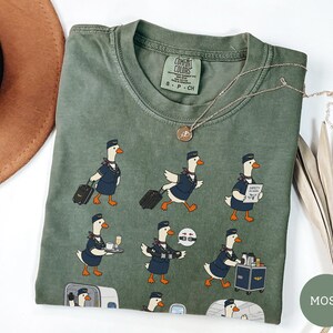 May include: A moss green crew neck t-shirt featuring a graphic print of cartoon ducks dressed as flight attendants performing various tasks, including pulling luggage, serving drinks, and demonstrating safety procedures on an aeroplane.
