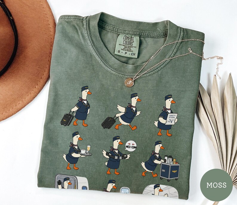 May include: A moss green crew neck t-shirt featuring a graphic print of cartoon ducks dressed as flight attendants performing various tasks, including pulling luggage, serving drinks, and demonstrating safety procedures on an aeroplane.