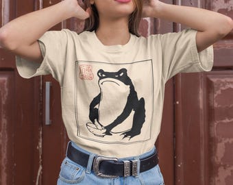 May include: A woman wearing a beige t-shirt with a black and white illustration of a frog holding a banana. The frog is in a square frame with a red seal in the top left corner. The woman is wearing a black hat and blue denim shorts.