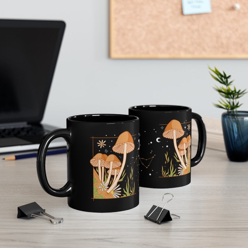 May include: Two black ceramic mugs with a mushroom design. The design features brown mushrooms, stars, and constellations on a black background. The mugs are on a light-colored wooden surface, with a laptop and office supplies in the background.