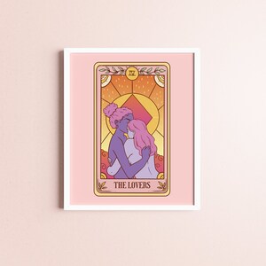 Op de afbeelding: Ingelijste kunstafdruk van een tarotkaart met twee figuren die elkaar omarmen, met de tekst "THE LOVERS". Het kunstwerk gebruikt een warm kleurenpalet met roze, oranje en paarse tinten. De afdruk is ingelijst met een witte rand.