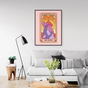 Op de afbeelding: Een ingelijste kunstdruk met een tarotkaartillustratie. De kaart, getiteld "THE LOVERS", toont twee figuren die elkaar omarmen, tegen een warme, zonsondergangkleurige achtergrond. Het kunstwerk is ingelijst in een zwart frame en hangt boven een grijze bank.