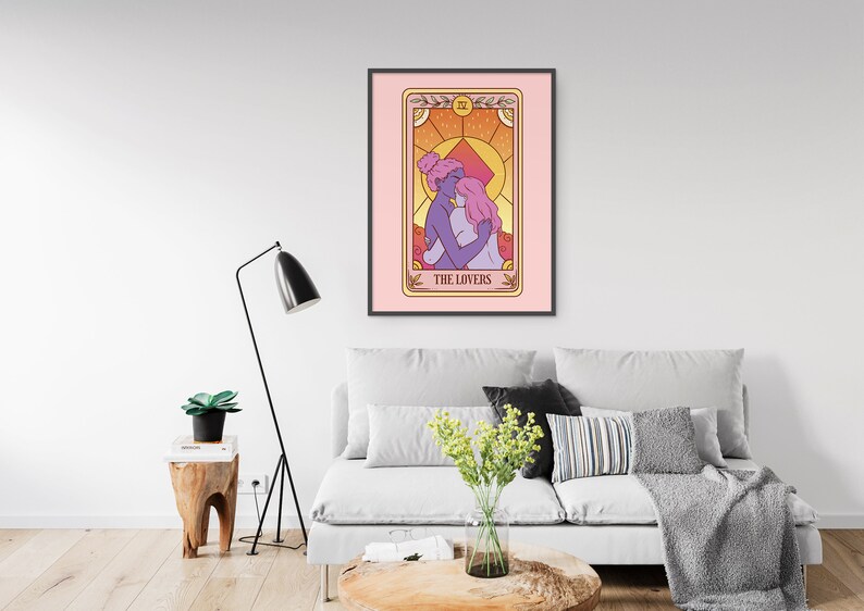 Op de afbeelding: Een ingelijste kunstdruk met een tarotkaartillustratie. De kaart, getiteld "THE LOVERS", toont twee figuren die elkaar omarmen, tegen een warme, zonsondergangkleurige achtergrond. Het kunstwerk is ingelijst in een zwart frame en hangt boven een grijze bank.