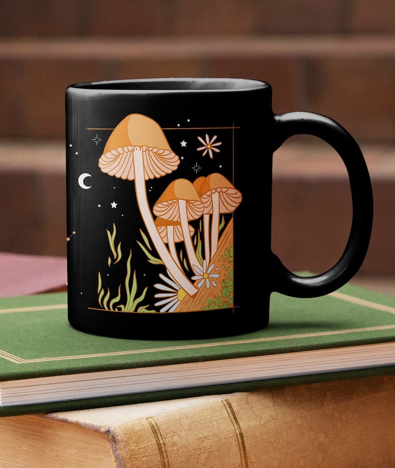 May include: Black ceramic mug featuring a whimsical illustration of orange mushrooms with white gills, a daisy, and stars against a night sky. The mug has a curved handle and is likely used for hot beverages.