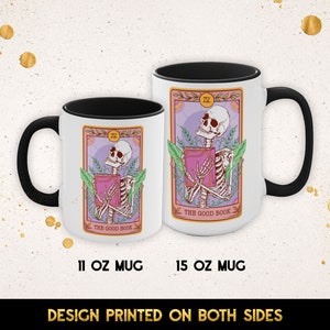 May include: Two white ceramic mugs with black handles. The mugs feature a colorful illustration of a skeleton holding a book with the text "The Good Book" and the Roman numeral "IV". The mugs are available in 11 oz and 15 oz sizes.