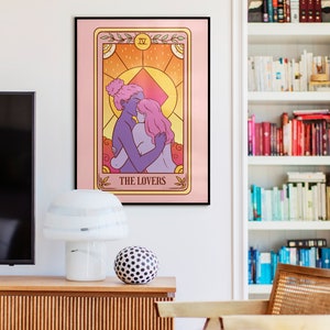 Op de afbeelding: Een ingelijste tarotkaartafdruk met een illustratie van twee figuren die elkaar omarmen, met de tekst "THE LOVERS" onderaan. Het kunstwerk heeft een roze achtergrond en een gouden rand. De afdruk hangt aan een muur boven een houten kast.
