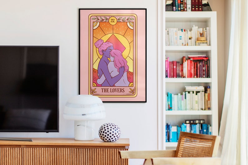 Op de afbeelding: Een ingelijste tarotkaartafdruk met een illustratie van twee figuren die elkaar omarmen, met de tekst "THE LOVERS" onderaan. Het kunstwerk heeft een roze achtergrond en een gouden rand. De afdruk hangt aan een muur boven een houten kast.