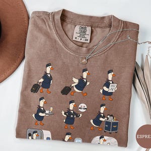 May include: An espresso-coloured t-shirt featuring a repeating graphic print of cartoon geese dressed as flight attendants. The geese are depicted performing various tasks like pulling luggage, serving drinks, and demonstrating safety procedures.