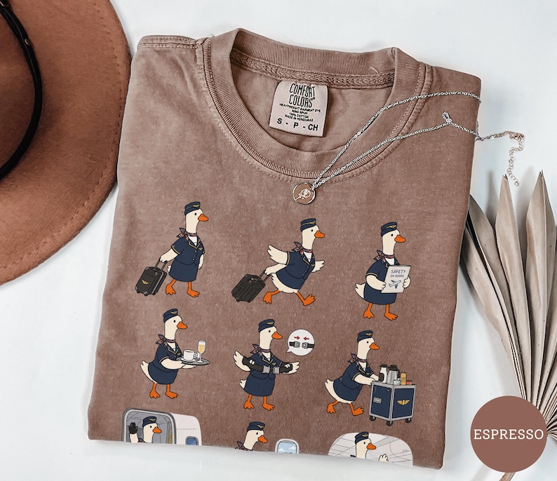 May include: An espresso-coloured t-shirt featuring a repeating graphic print of cartoon geese dressed as flight attendants. The geese are depicted performing various tasks like pulling luggage, serving drinks, and demonstrating safety procedures.
