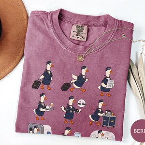 May include: A berry-coloured crew neck t-shirt featuring a repeating graphic print of white geese dressed as flight attendants in navy blue uniforms. The geese are depicted performing various tasks like pulling luggage, serving drinks, and demonstrating safety.