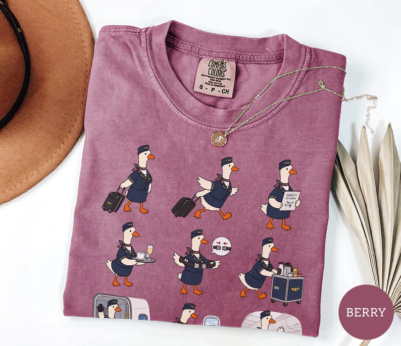 May include: A berry-coloured crew neck t-shirt featuring a repeating graphic print of white geese dressed as flight attendants in navy blue uniforms. The geese are depicted performing various tasks like pulling luggage, serving drinks, and demonstrating safety.