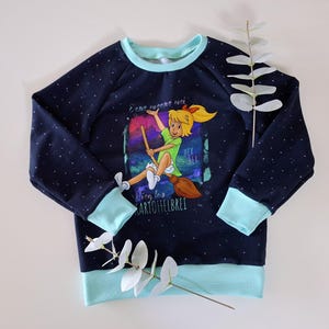 Könnte beinhalten: Dunkelblaues Sweatshirt mit hellblauen Bündchen und Saum. Auf der Vorderseite befindet sich ein farbenfrohes Motiv einer Cartoon-Hexe auf einem Besen, mit dem Text "Flieg los Kartoffelbrei". Das Sweatshirt hat ein Polka-Dot-Muster.