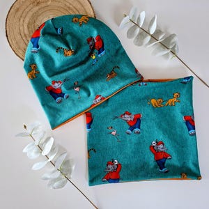 Könnte beinhalten: Ein tealfarbenes Mützen- und Halstuch-Set mit einem sich wiederholenden Cartoon-Elefanten-, Löwen- und Tigermuster. Das Set hat eine orangefarbene Einfassung und ist aus einem weichen Stoff gefertigt. Die Mütze ist umgeschlagen, und das Halstuch ist flach.