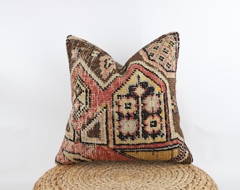 May include: A square throw cushion featuring a woven textile cover with a complex geometric pattern in shades of brown, red, yellow, and cream. The design includes repeating angular shapes, diamonds, and floral-like motifs across the surface.
