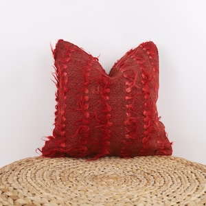 Könnte beinhalten: Ein rotes Zierkissen mit einem strukturierten, zotteligen Design. Das Kissen ist rechteckig und liegt auf einem gewebten, kreisförmigen Ottomanen. Die Farbe des Kissens ist ein tiefes, leuchtendes Rot und verleiht jedem Raum einen Farbtupfer.