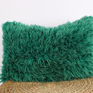May include: A soft, plush pillow with a deep green, fuzzy texture. The pillow is rectangular and has a slightly rounded shape. It is sitting on a woven, straw-like surface.