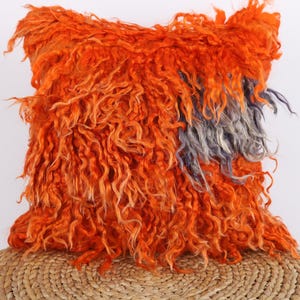 Peut inclure: Un coussin carré orange avec une surface texturée et hirsute. Un morceau de fibres grises et bleues est visible sur le côté droit du coussin. Le coussin repose sur une surface tressée de couleur beige.