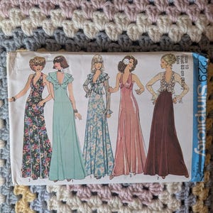 Op de afbeelding: Vintage Simplicity naaipatroon met illustraties van vijf lange jurken met verschillende halslijnen en mouwstijlen. De jurken zijn afgebeeld in bloemen-, effen en bedrukte stoffen. Het patroon is gelabeld "2290 Simplicity" en bevat maatdetails.