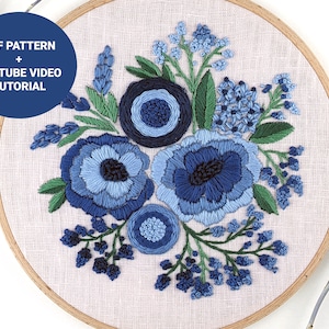 May include: Embroidered hoop art featuring a blue floral design with a variety of blue flowers and leaves. The hoop is made of wood and has a white linen fabric background. The text "PDF PATTERN + YOUTUBE VIDEO TUTORIAL" is displayed in a blue circle.