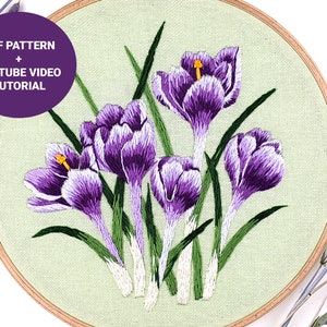 May include: Embroidered hoop art featuring a cluster of purple crocuses with green stems and leaves. The hoop is made of wood and has a light green fabric background. The text "PDF PATTERN + YOUTUBE VIDEO TUTORIAL" is displayed in a purple circle.