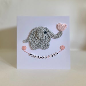 Puede incluir: Un elefante de crochet gris sosteniendo un coraz&oacute;n de crochet rosa. El elefante est&aacute; en una tarjeta blanca con las palabras "WELCOME ISLA" escritas en cuentas rosas y blancas.