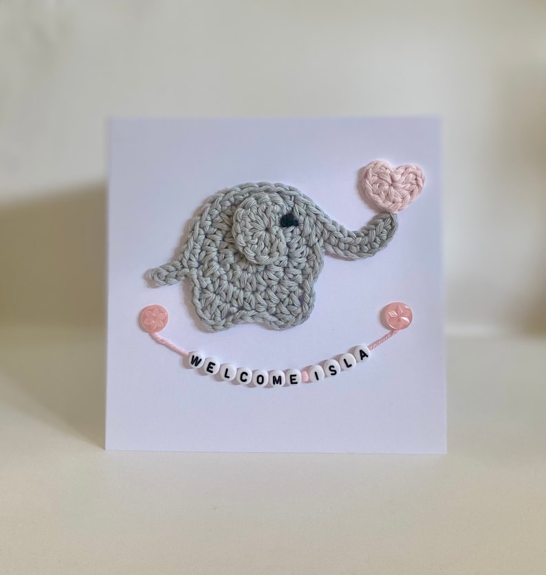 Puede incluir: Un elefante de crochet gris sosteniendo un coraz&oacute;n de crochet rosa. El elefante est&aacute; en una tarjeta blanca con las palabras "WELCOME ISLA" escritas en cuentas rosas y blancas.