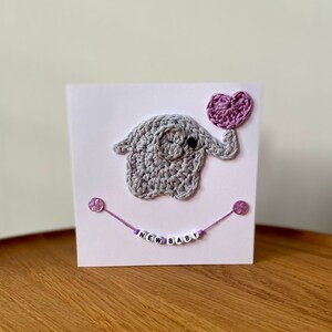 Puede incluir: Un elefante de crochet gris sosteniendo un globo de coraz&oacute;n morado. El elefante est&aacute; en una tarjeta blanca con una cuerda morada y cuentas que deletrean "NEW BABY".