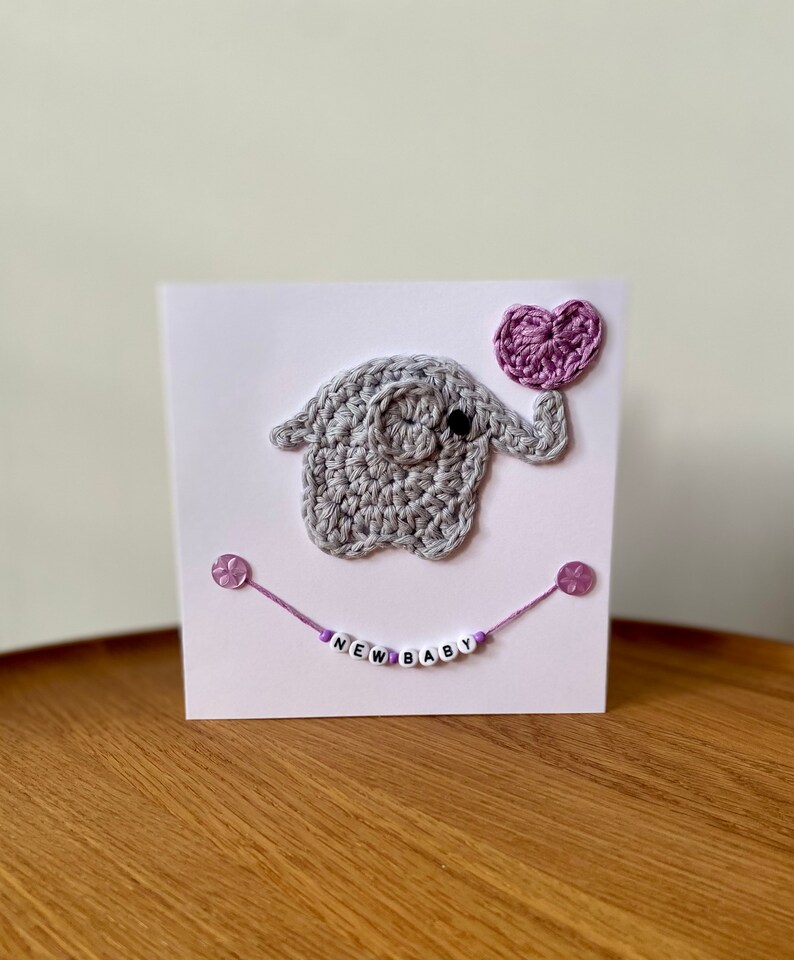Puede incluir: Un elefante de crochet gris sosteniendo un globo de coraz&oacute;n morado. El elefante est&aacute; en una tarjeta blanca con una cuerda morada y cuentas que deletrean "NEW BABY".