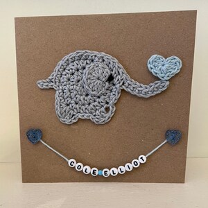 Puede incluir: Un elefante de crochet gris que sostiene un coraz&oacute;n de crochet azul claro. El elefante est&aacute; en una tarjeta marr&oacute;n con una cadena de cuentas blancas con el nombre "COLE ELLIOT" escrito en letras negras.