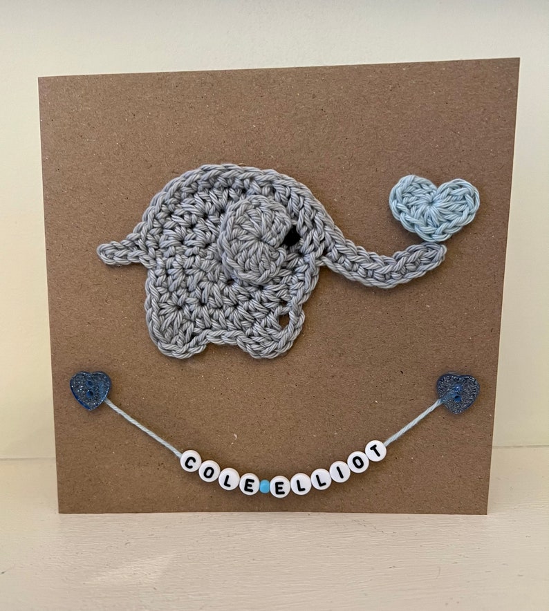 Puede incluir: Un elefante de crochet gris que sostiene un coraz&oacute;n de crochet azul claro. El elefante est&aacute; en una tarjeta marr&oacute;n con una cadena de cuentas blancas con el nombre "COLE ELLIOT" escrito en letras negras.