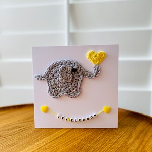 Puede incluir: Un elefante de crochet gris que sostiene un coraz&oacute;n de crochet amarillo. El elefante est&aacute; en una tarjeta blanca con una cadena de cuentas amarillas que deletrean "IVY ELEANOR".